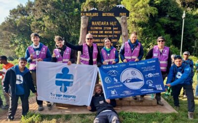 A group of hikers, some in purple vests, poses with flags in front of a wooden sign for Mandara Hut in Kilimanjaro National Park. One person is lying on the ground in front, holding a flag.